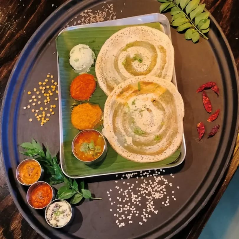 Two dosas with various chutneys and sambar, garnished with curry leaves and spices on a dark tray.