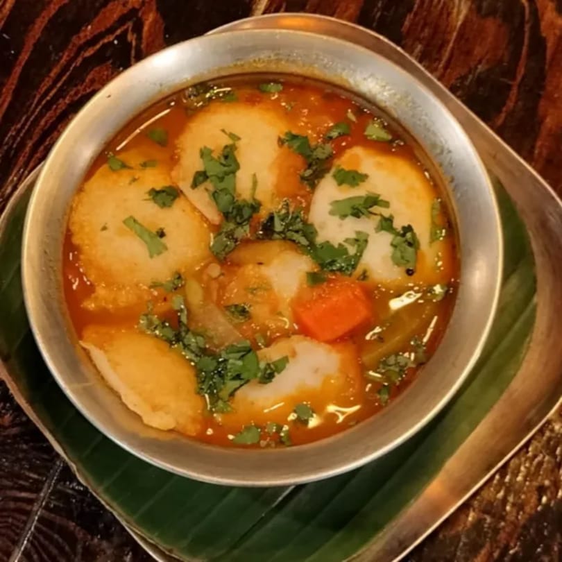 A metal bowl of Indian sambar with idli, vegetables, and cilantro, served on a banana leaf.